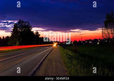 Sentieri leggeri che partono dalle auto di passaggio sulla strada rurale al tramonto con il colorato cielo serale Foto Stock