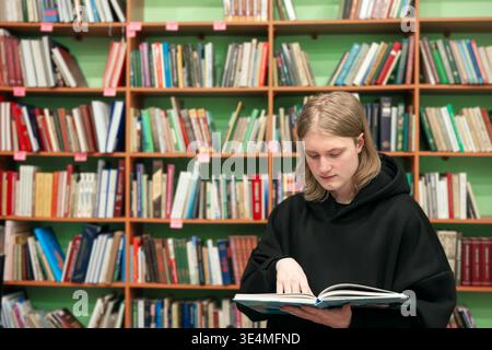 Giovane che legge un libro in una biblioteca pubblica. Studentessa bionda intelligente in biblioteca, concetto educativo. Lettura, apprendimento e ricerca accademica, acquisto di textboo Foto Stock