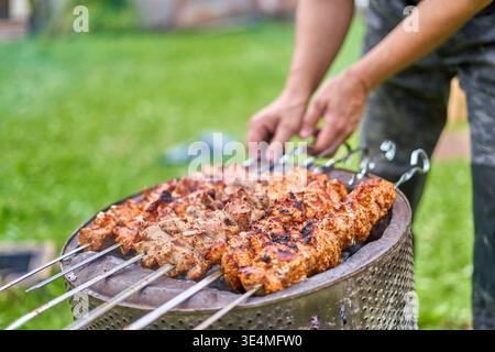 Vista ravvicinata di più spiedini di carne marinati che grigliano su un barbecue di metallo rotondo in un ambiente esterno. Diversi spiedini con carne condita Foto Stock