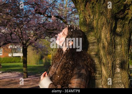 Giovane donna in piedi vicino a un albero sotto fiori di ciliegio in un parco. Luce solare calda, stagione primaverile e atmosfera tranquilla all'aperto. Foto Stock