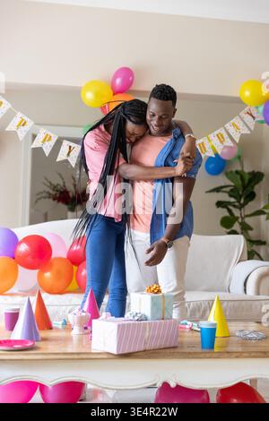 Coppia afroamericana che abbraccia a casa, festeggia il compleanno con striscioni di compleanno e palloncini Foto Stock
