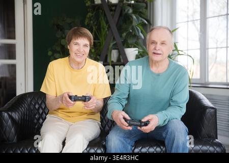 si sta godendo una battaglia virtuale. Concetto di Home Entertainment. Una coppia felice di anziani gioca ai videogame con le console insieme a casa sedute sul divano || modello rilasciato Foto Stock