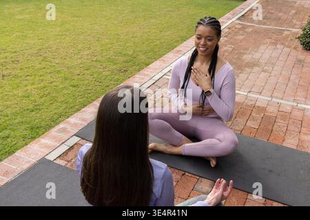 Donne diverse che respirano su tappeti su traiettorie di mattoni in erba, indossano smartwatch, abbigliamento sportivo, spazio per copiare. All'aperto, yoga, istruttore, meditazione, gambe incrociate, lavanda, viola || modello rilasciato Foto Stock