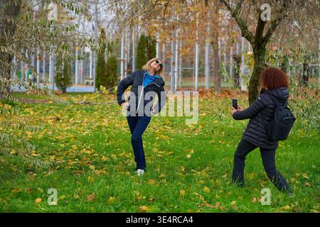 Donna di mezza età che scatta una foto su smartphone della sua amica bionda in posa nel parco autunnale Foto Stock