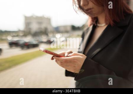 Donna dai capelli rossi sicura che usa il telefono cellulare all'aperto. Look elegante, atmosfera cittadina, giorno di sole, tecnologia e concetto di comunicazione. Giovane donna adulta f Foto Stock