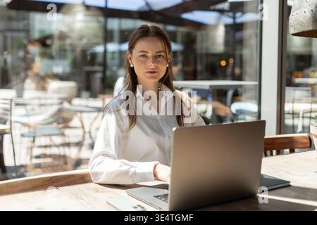 Libero professionista che scrive sul laptop in un accogliente bar. Area di lavoro moderna con caffè, telefono e accessori eleganti. Ambiente di lavoro remoto produttivo. Donna che lavora Foto Stock