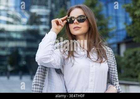 Elegante giovane professionista che indossa occhiali da sole e abiti eleganti all'aperto nel quartiere finanziario. Donna elegante con occhiali da sole che posa con fiducia Foto Stock
