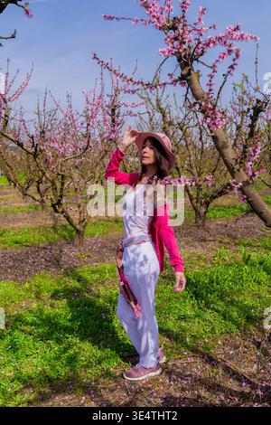 Donna in piedi in un vivace frutteto di pesca durante la primavera, godendo della bellezza della natura circondata da delicati fiori rosa e erba verde sotto Foto Stock