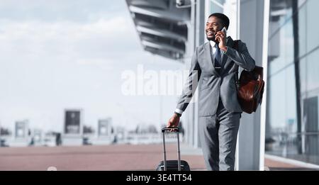 Uomo d'affari afroamericano che cammina con i bagagli e parla al telefono Foto Stock