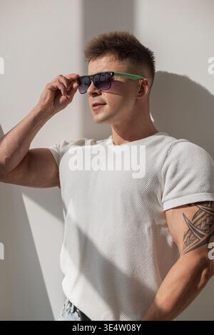 Giovane uomo elegante con occhiali da sole che indossa con sicurezza un outfit casual: T-shirt bianca e jeans strappati. Ambiente interno moderno e minimale con solarium naturale Foto Stock