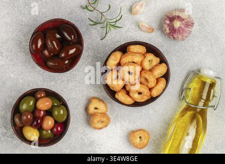 Cibo, Tarallini: Piccoli bagel italiani tradizionali. Taralli. Snack gustoso. Olive nere e verdi, Kalamata, olio d'oliva, aglio e rosmarino su un condimento grigio Foto Stock