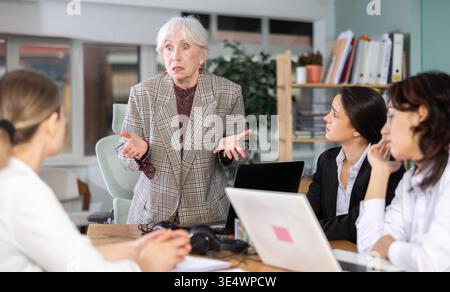 Donna anziana in piedi e rimprovera colleghi seduti tristi in ufficio Foto Stock