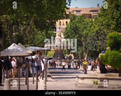 AIX-en-Provence, Francia - 2 luglio 2024: I turisti e la gente del posto si godono una giornata di sole nella storica piazza della città con fontana ornata e bancarelle del mercato Foto Stock