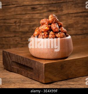 Arachidi arrostite caramellate servite in una ciotola di legno, con una consistenza croccante e una lucentezza dolce, su uno sfondo rustico - Arachis ipogaea Foto Stock