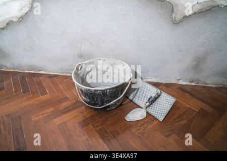 Utensili per intonacare su un pavimento in legno contro una parete in cemento non finita Foto Stock
