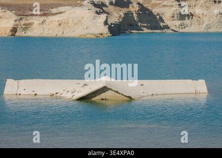 Una vista più ravvicinata di una casa sul tetto in cemento parzialmente sommersa dalle acque cristalline turchesi di un lago di cava. La struttura mostra segni di tempo Foto Stock