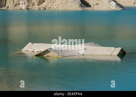 Una vista più ravvicinata di una casa sul tetto in cemento parzialmente sommersa dalle acque cristalline turchesi di un lago di cava. La struttura mostra segni di tempo Foto Stock