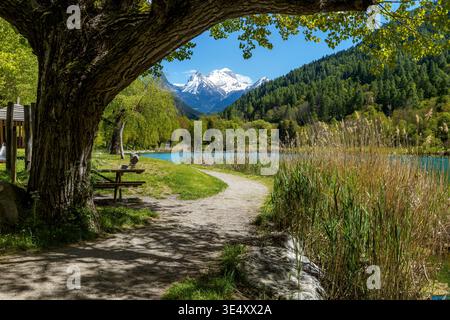 Una vista pittoresca di un piccolo lago Lauzet-Ubaye e di un parco naturale, caratterizzato da alberi lussureggianti e sentieri con una montagna innevata sullo sfondo. Foto Stock