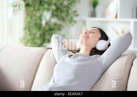 Felice donna asiatica che indossa cuffie wireless e si rilassa seduto sul divano di casa Foto Stock