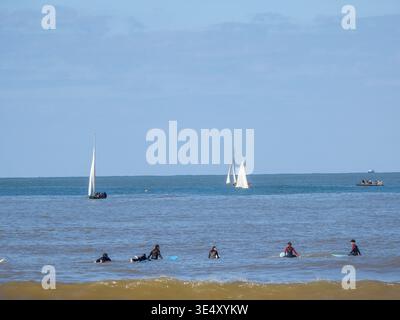 I surfisti in muta sono in attesa di onde sulle loro tavole vicino alla riva, mentre le barche a vela e un gommone con i passeggeri attraversano il mare blu sotto una Foto Stock
