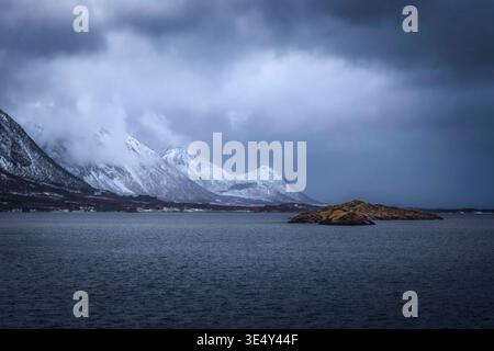Le vette innevate si innalzano maestosamente sulle acque scure di Fiskebol, Lofoten, Norvegia. Un freddo riempie l'aria mentre le nuvole girano sopra, creando un'atmosfera Foto Stock