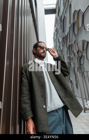 Uomo elegante con abiti alla moda in piedi vicino al muro e indossare occhiali da sole in città. Ragazzo hipster urbano alla moda Foto Stock