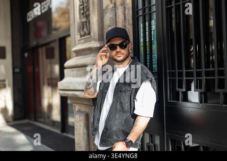 Un uomo alla moda elegante e alla moda indossa occhiali da sole in berretto nero con gilet in denim e stativi bianchi per T-shirt nella città di Barcellona Foto Stock
