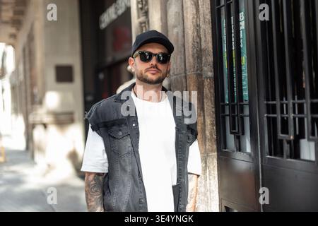 Elegante e alla moda, uomo hipster con occhiali da sole in berretto nero con gilet in denim e T-shirt bianca che cammina nella città di Barcellona Foto Stock