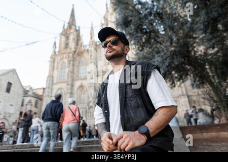 Viaggiatore uomo elegante e affascinante con occhiali da sole con cappello e giubbotto in denim che viaggia a Barcellona vicino alla chiesa d'epoca Foto Stock