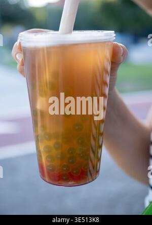 Primo piano di una mano che regge una tazza trasparente riempita di colorato tè con perle di tapioca e un'ampia paglia su uno sfondo esterno Foto Stock