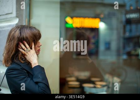 La figura femminile con i capelli marroni che indossano una giacca nera si erge fuori da un bar, toccando le orecchie mentre si riflette nella finestra di vetro con i tavoli visibili Foto Stock