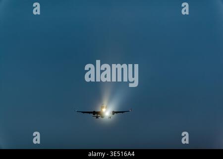 Aereo in avvicinamento all'atterraggio con luci di atterraggio illuminate contro un cielo blu scuro durante il crepuscolo, che mostrano la silhouette dell'aereo e. Foto Stock