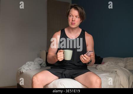 Un uomo caucasico di 30 anni si siede sul bordo di un letto non fatto, tenendo in mano una tazza di caffè verde e uno smartphone. La scenografia presenta uno sfondo neutro Foto Stock