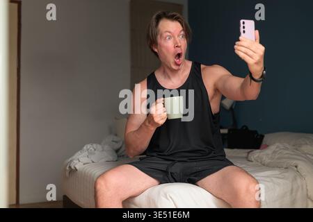 Un uomo caucasico medio adulto si siede su un letto con un'espressione scioccata mentre guarda il suo smartphone e tiene in mano una tazza. Il design minimalista per interni Foto Stock