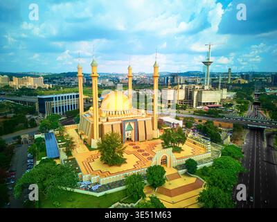 Vista aerea di una moschea dalla cupola dorata circondata da vegetazione lussureggiante e architettura moderna sotto un vasto cielo nuvoloso, Abuja, Nigeria. Foto Stock
