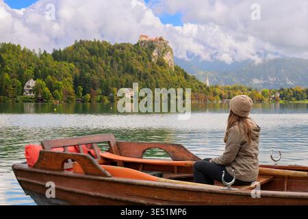 Una giovane donna siede su una barca a remi di legno sul lago di Bled, con il castello medievale di Bled in lontananza all'inizio dell'autunno nelle Alpi Giulie a Bled in Slovenia Foto Stock
