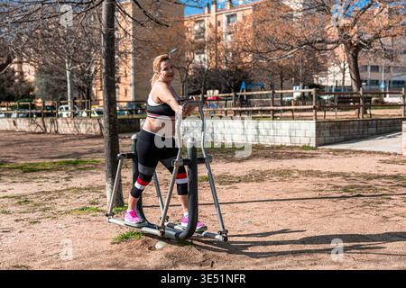 Donna anziana impegnata in attività fitness all'aperto, felicemente utilizzando una macchina ellittica in un parco pubblico sotto un cielo azzurro e limpido, promuovendo un'aria condizionata Foto Stock