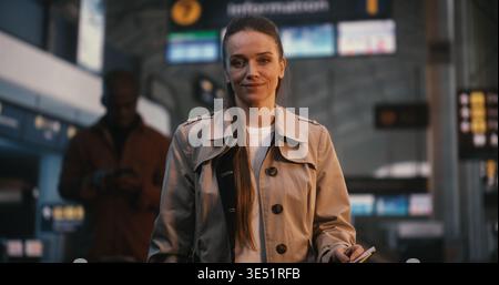 Il ritratto della splendida giovane donna con sorriso si trova nel vivace terminal dell'aeroporto. Vestita con un classico Trench beige femminile che esprime entusiasmo e gioia per il prossimo viaggio. Donna di stile. Foto Stock