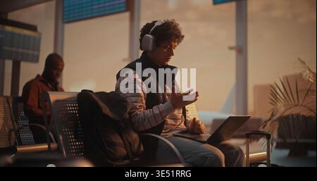 Giovane professionista che effettua una videochiamata aziendale all'aeroporto con l'aereo in partenza alle spalle. Giovane uomo con capelli ricci partecipa a importanti riunioni di lavoro tramite laptop. Concetto di comunicazione globale. Foto Stock