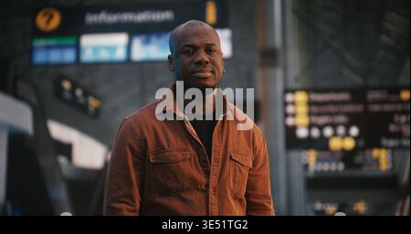 Maschio afroamericano si erge con sicurezza e sorriso sottile nell'area partenze dell'aeroporto internazionale. Terminal dell'ambiente vivace, con le persone che camminano, in evidenza il comportamento incentrato sull'uomo. Verticale. Foto Stock
