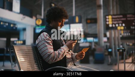 Seduto nella lounge dell'aeroporto, Young Man with Curly Hair utilizza lo smartphone per eseguire la scansione del pass d'imbarco digitale, resta concentrato sullo schermo, scorrendo i dettagli del volo, il numero del gate e l'orario di partenza. Foto Stock