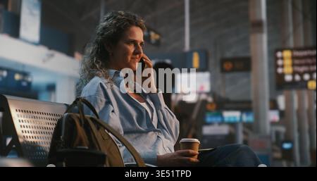 Una giovane donna siede comodamente nella lounge dell'aeroporto, gustando un caffè mentre si fa una piacevole conversazione telefonica. Esperienza di viaggio senza stress, emozioni positive e restare in contatto con i propri cari. Foto Stock