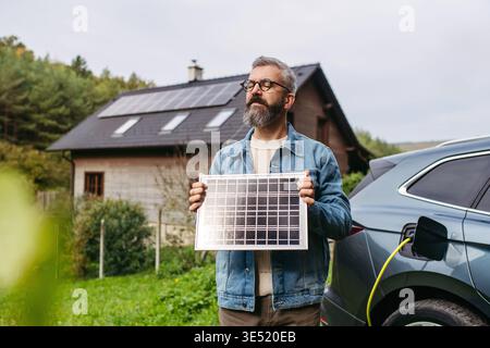 Uomo che tiene in mano il modello di pannello solare. Ricarica auto elettrica e casa con sistema a pannelli solari sul tetto dietro di lui. Foto Stock