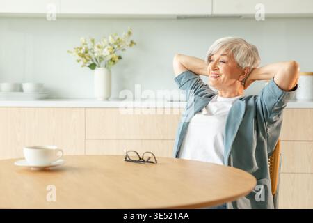 Una donna anziana si sente rilassata e si allunga le braccia in cucina a casa. Una donna matura sorridente che si allunga le mani si sente positiva ottimista fresca nel nuovo giorno della mattina. Buon umore felice stile di vita anziano Foto Stock