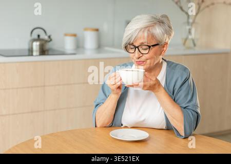 Buon giorno. Comfort rilassato anziano anziano anziano anziano donna nonna che beve bevande calde tè e caffè in cucina a casa. Donna matura che riposa goditi la calma mattinata nella casa moderna da sola. Concetto di pensione Foto Stock
