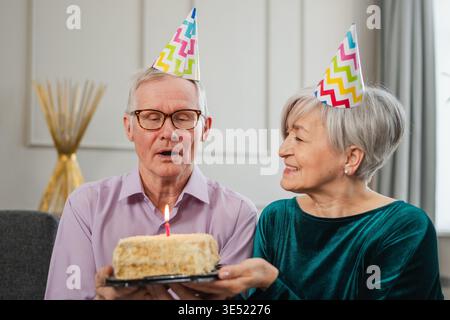 Esprimi un desiderio. La coppia senior della famiglia in cappello per festeggiare l'anniversario del compleanno insieme a casa. Vecchio che fa saltare le candele bruciate sulla torta di compleanno. La vecchia donna augura buon compleanno al marito Foto Stock