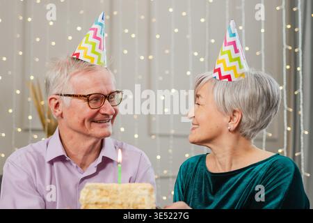Esprimi un desiderio. La coppia senior della famiglia in cappello per festeggiare l'anniversario del compleanno insieme a casa. Una vecchia donna che spara candele bruciate sulla torta di compleanno. Il vecchio augura alla moglie buon compleanno Foto Stock