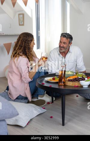 Coppia seduta a casa sui cuscini intorno al tavolino da caffè, condividendo vino, tagliere di formaggi, petali di rosa Foto Stock