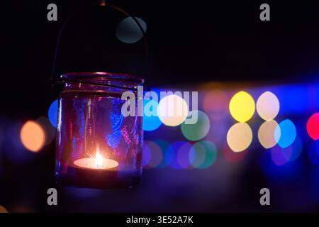 Una fotografia ravvicinata di una candela illuminata che brucia all'interno di una lanterna in vetro trasparente di colore viola. La lanterna è appesa a un metallo curvo Foto Stock