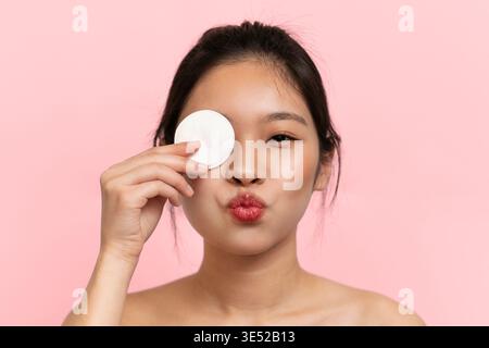 Una giovane donna tiene il tampone di cotone al suo occhio mentre crea un volto giocoso su sfondo rosa Foto Stock